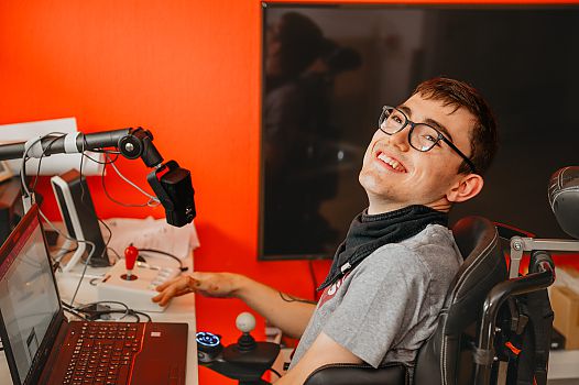 Ein junger Mann mit E-Rolli lacht. Er bedient einen Laptop. Neben ihm ist ein großes Mikrofon.